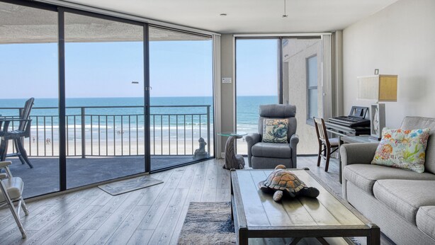 Una habitación con vista a la playa, un ventilador de techo, un piano y una tortuga sobre una mesa de centro.