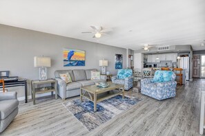 A modern living room with a gray sofa, blue patterned chairs, a wooden coffee table, and a kitchen in the background.