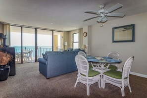 A room with a ceiling fan, a dining table with chairs, a sofa, and a view of the ocean through large windows.