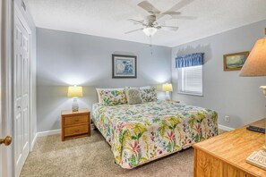 Une chambre à coucher comprenant un lit, deux tables de chevet avec des lampes, un ventilateur de plafond, une fenêtre avec des stores et une porte.