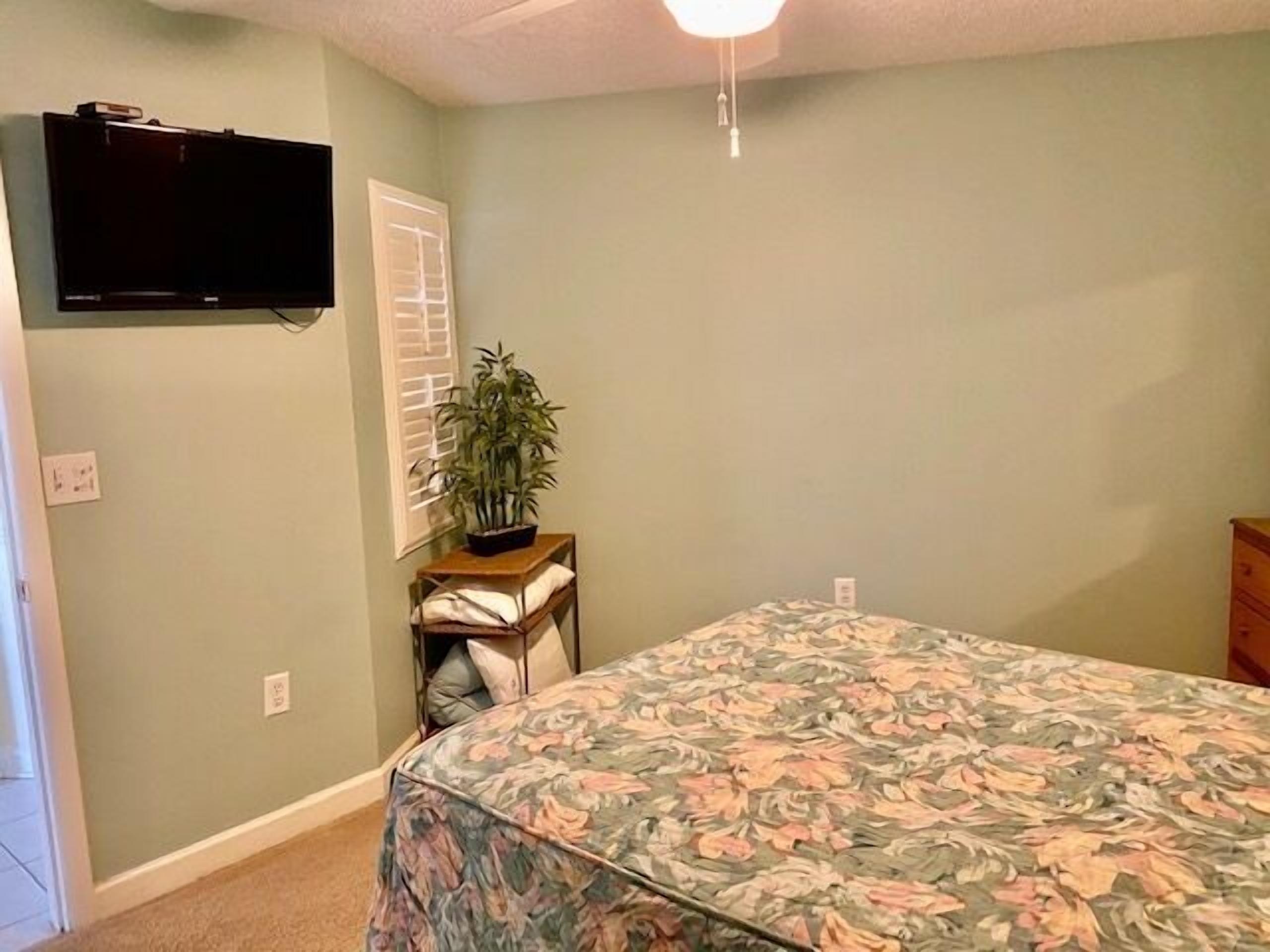 A bedroom with a bed, a television mounted on the wall, a window with blinds, a small table with a plant, and a nightstand.