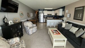Sala de estar com sofá, poltrona e mesa de centro. Há uma cozinha com geladeira e micro-ondas.