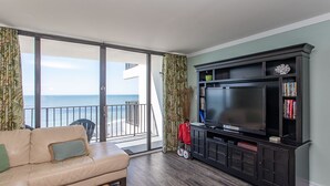 A living room with a sofa, a TV, and a bookshelf. There is a view of the ocean through large glass doors. - Immaculate, flawless two bedroom condo (Murrells Inlet)