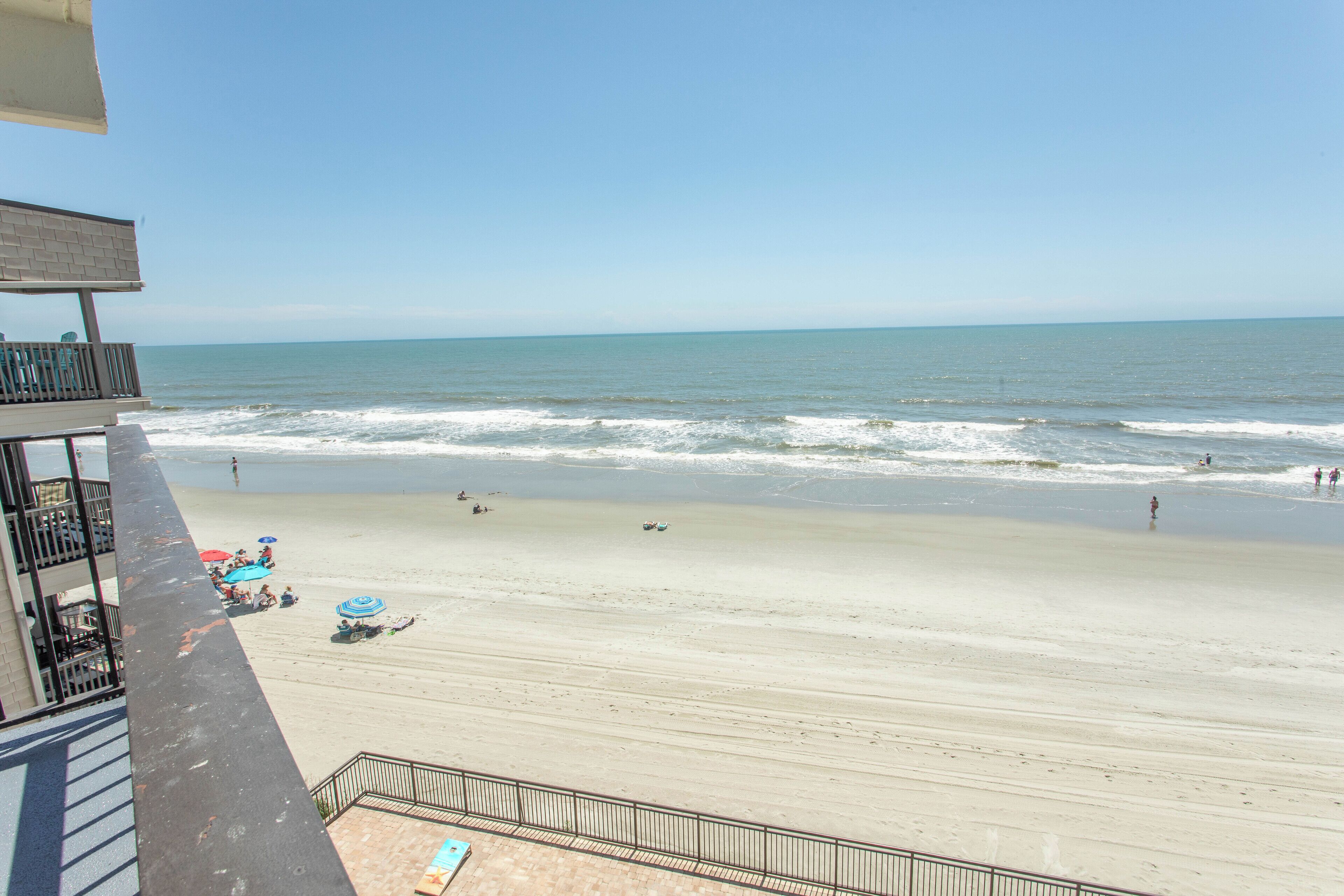 A beach view from a balcony with a railing, a sandy beach, and the ocean.