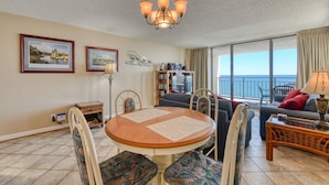 A living room with a round table, chairs, a sofa, a balcony with a view of the sea, and a bookshelf. - Surfmaster 909 - One-bedroom, one-and-a-half-bath oceanfront condo with pools (Garden City Beach)