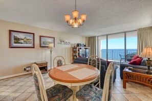 A living room with a round table, chairs, a sofa, a balcony with a view of the sea, and a bookshelf.