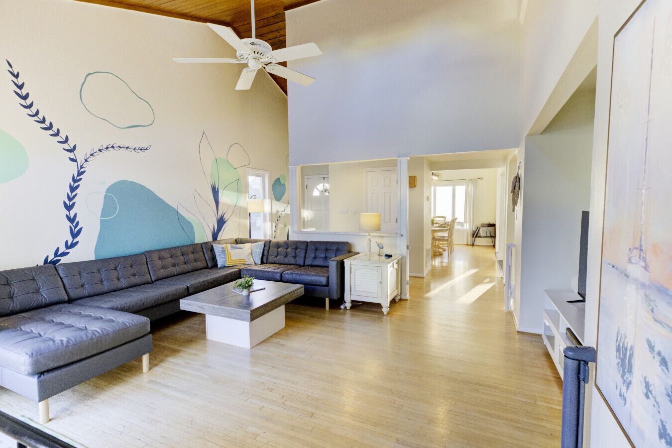 A spacious living room with a grey sectional sofa, a coffee table, and a white cabinet. There is a ceiling fan, a painting on the wall, and a view into the dining area.