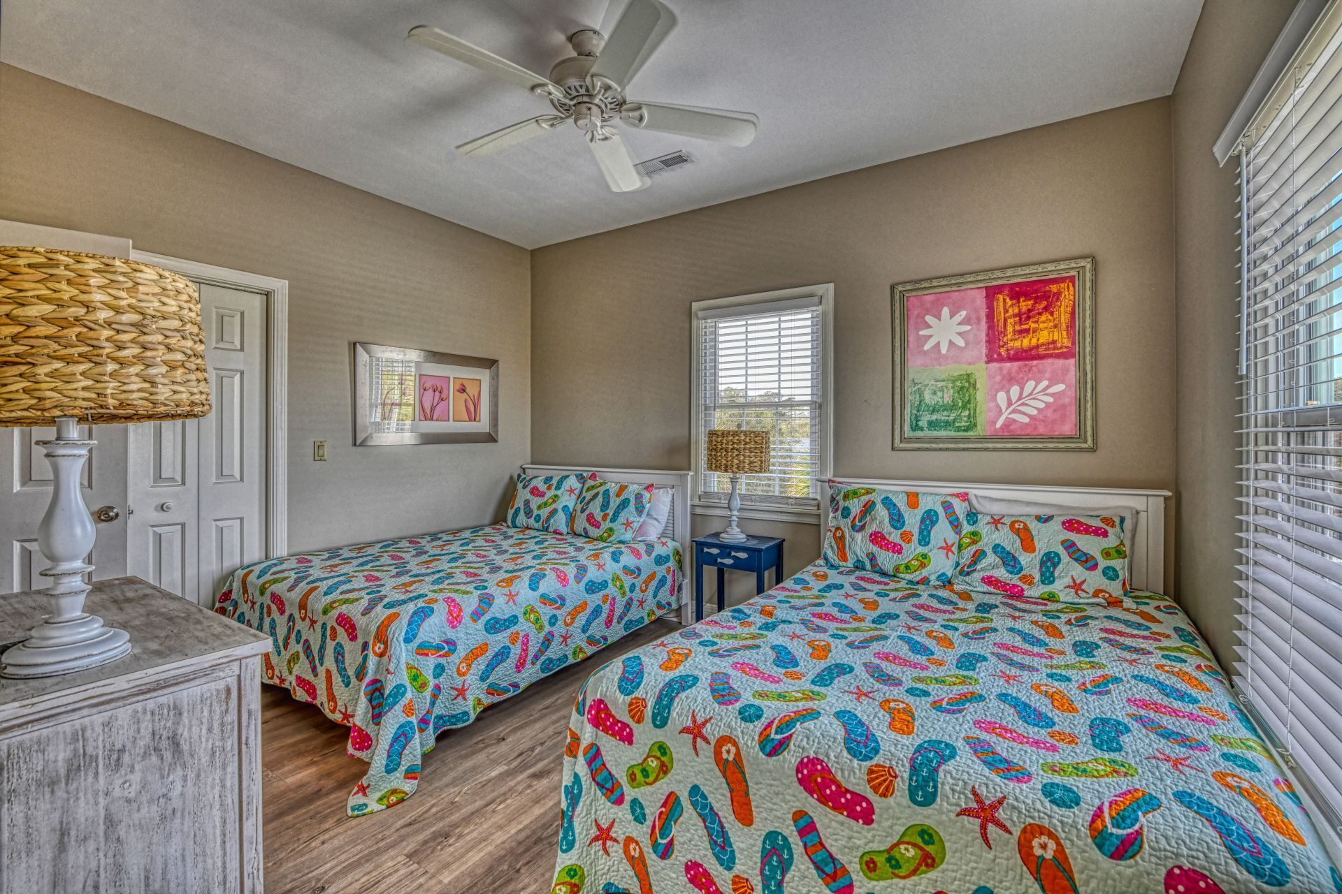 A bedroom with two beds, a ceiling fan, a lamp, and artwork on the walls.