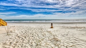 Ein Strand mit einer Holztreppe, die zum Sand hinunterführt, einem Holzpfahl im Sand und dem Meer im Hintergrund.