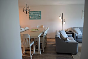 A dining area with a wooden table and white chairs, a gray sofa, and a wall-mounted light fixture.