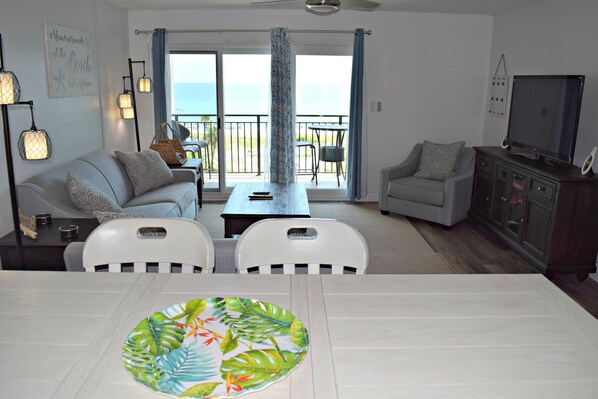 A living room with a white table, two white chairs, a grey sofa, a grey armchair, a TV, and a balcony with a view of the ocean.