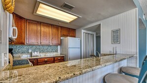 Una cocina con armarios de madera, encimera de granito, refrigerador y una silla azul.