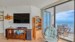 Sala de estar con televisor de pantalla plana, mueble de entretenimiento de madera y un sillón cómodo junto a un amplio ventanal corredera que da a un balcón con vistas al océano.
