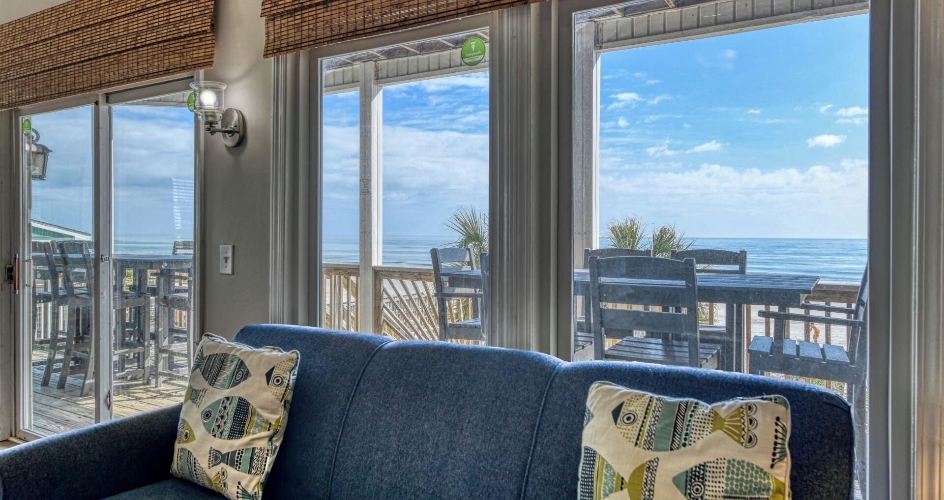 A room with a view of the ocean, a blue sofa with patterned cushions, and a wooden deck with outdoor seating.