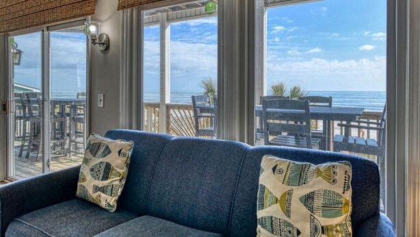 Et værelse med en blå sofa, mønstrede puder og store vinduer med udsigt over en strand.