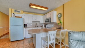 Una cocina compacta con armarios blancos, un refrigerador, un microondas y un lavamanos. Hay una pequeña zona de comedor con una silla blanca y un sofá.