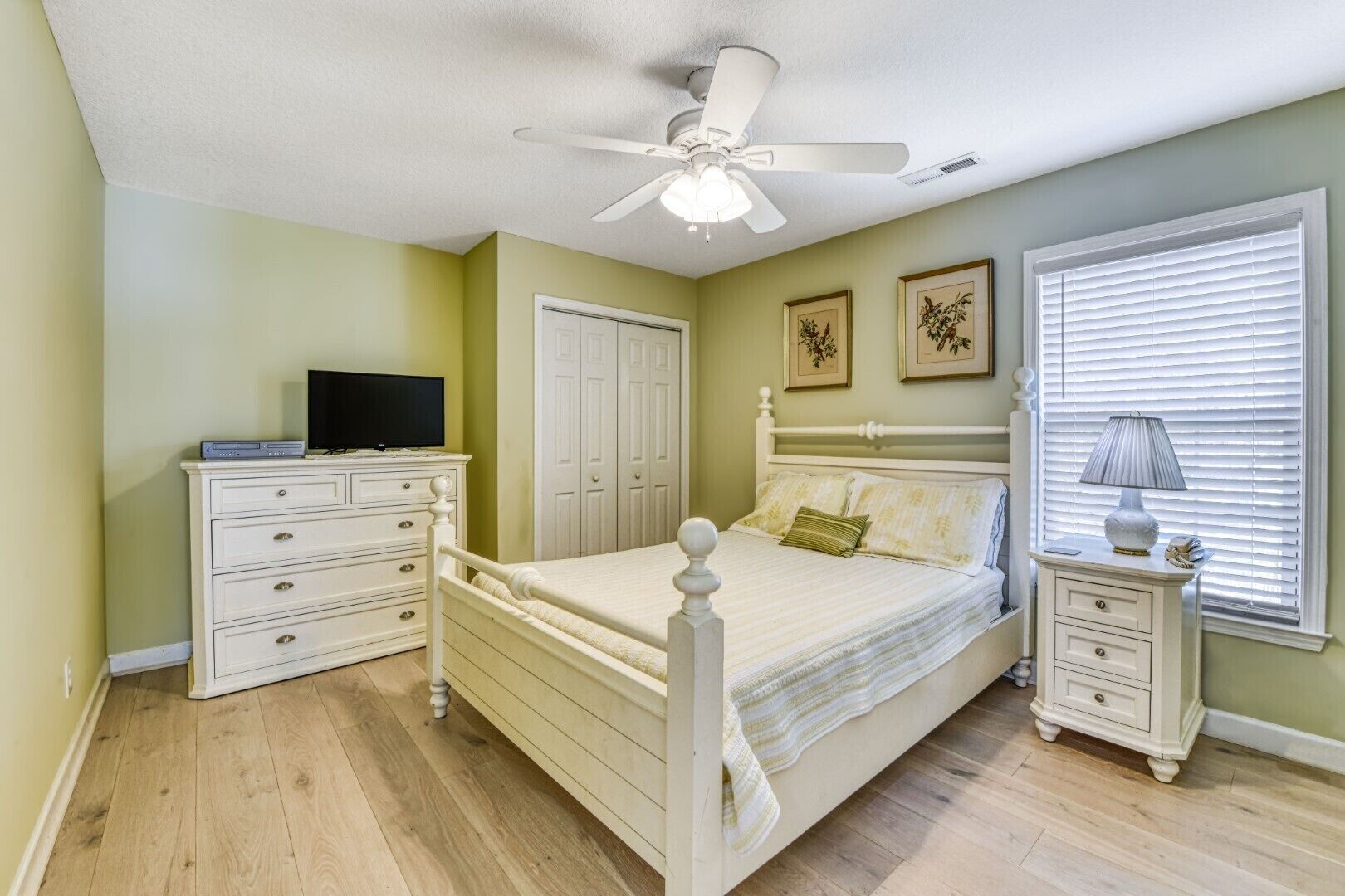A bedroom with a bed, dresser, nightstand, ceiling fan, and a window with blinds.