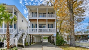 A two-story beachfront property with a balcony, a covered porch, and a staircase leading to the upper floor.