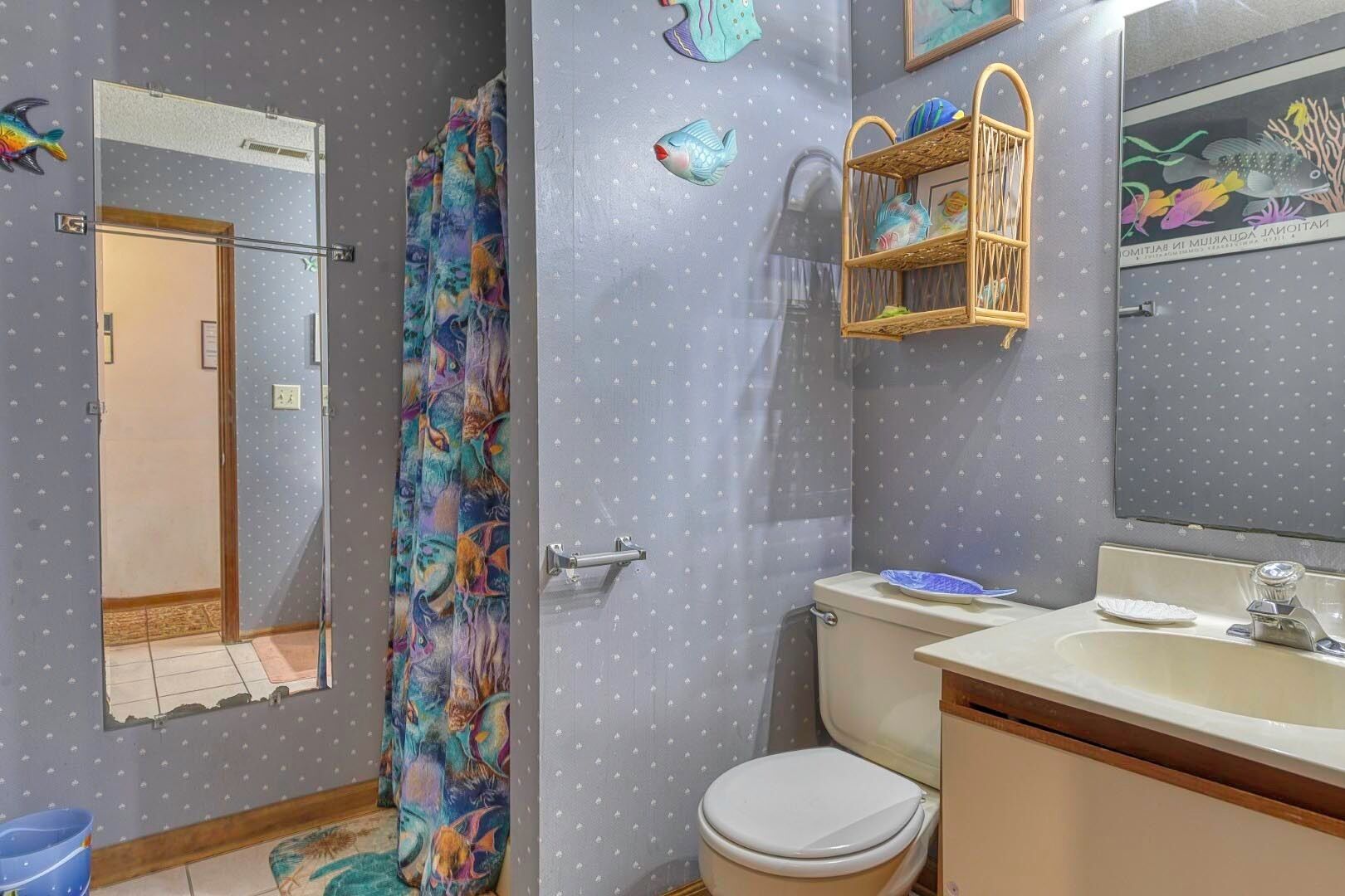 A bathroom with a shower curtain featuring a colorful fish pattern, a toilet, a sink with a mirror above it, and a small shelf with decorative items.