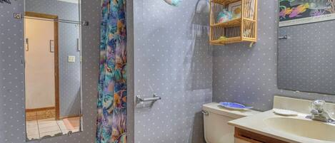 A bathroom with a shower curtain featuring a colorful fish pattern, a toilet, a sink with a mirror above it, and a small shelf with decorative items.