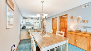 A dining area with a wooden table and chairs, a chandelier, and a kitchen in the background. - Sandy Shores II 209 - Two-bedroom, two-bath oceanview condo in GCB (Garden City Beach)