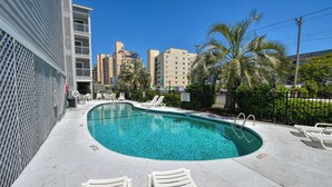 A swimming pool with lounge chairs, surrounded by a fence and palm trees, with buildings in the background. - Sandy Shores II 209 - Two-bedroom, two-bath oceanview condo in GCB (Garden City Beach)