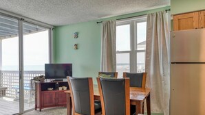 A room with a sliding glass door leading to a balcony, a wooden dining table with chairs, a television on a cabinet, and a refrigerator. - Sea Watch VIII 103 - Two Bedroom, One Bath Oceanfront Condo (Garden City Beach)