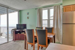 A room with a sliding glass door leading to a balcony, a wooden dining table with chairs, a television on a cabinet, and a refrigerator.
