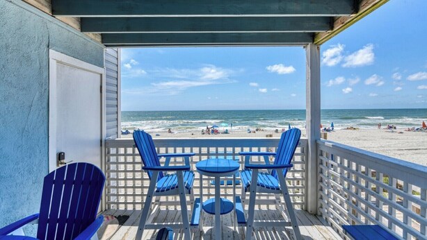 A beachside balcony with blue Adirondack chairs and a table, overlooking a sandy beach with people and umbrellas. - Sea Watch VIII 103 - Two Bedroom, One Bath Oceanfront Condo (Garden City Beach)