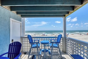A beachside balcony with blue Adirondack chairs and a table, overlooking a sandy beach with people and umbrellas.