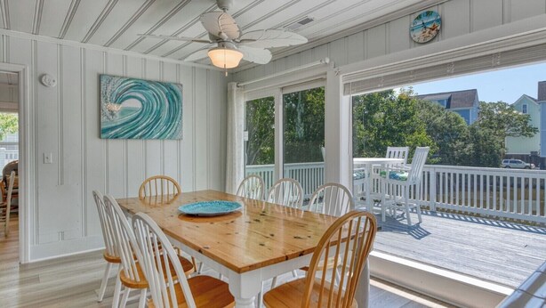 A dining area with a wooden table and white chairs, a ceiling fan, and a view of the deck and greenery outside. - Charming Coastal - 5 Bedroom, 4 Bath Beach House with Private Pool (Garden City Beach)