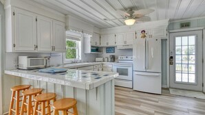 A kitchen with white cabinets, a central island with stools, a ceiling fan, and a view of the outside through a window. - Charming Coastal - 5 Bedroom, 4 Bath Beach House with Private Pool (Garden City Beach)