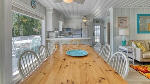 A spacious dining area with a wooden table and white chairs, a kitchen with white cabinets, and a view of the outdoors. - Charming Coastal - 5 Bedroom, 4 Bath Beach House with Private Pool (Garden City Beach)