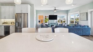 A modern kitchen with white cabinets, a stainless steel refrigerator, and a dining area with a white table and chairs. There are three pendant lights hanging above the table.