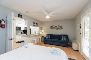 Une chambre d’hôtel avec un lit, un canapé bleu foncé, un petit coin cuisine et un ventilateur de plafond.