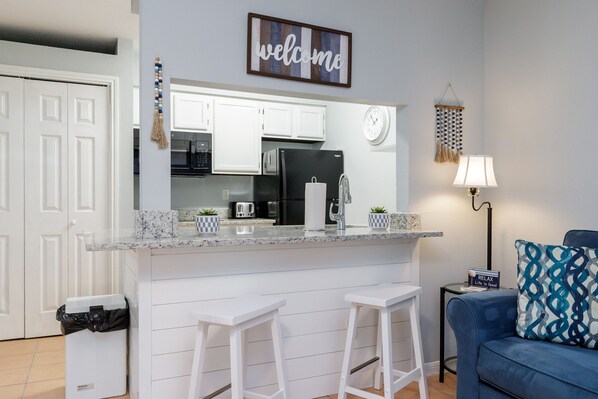 A modern kitchen with white cabinets, a black refrigerator, and a welcome sign above the counter. There are two white bar stools and a blue couch with patterned cushions.
