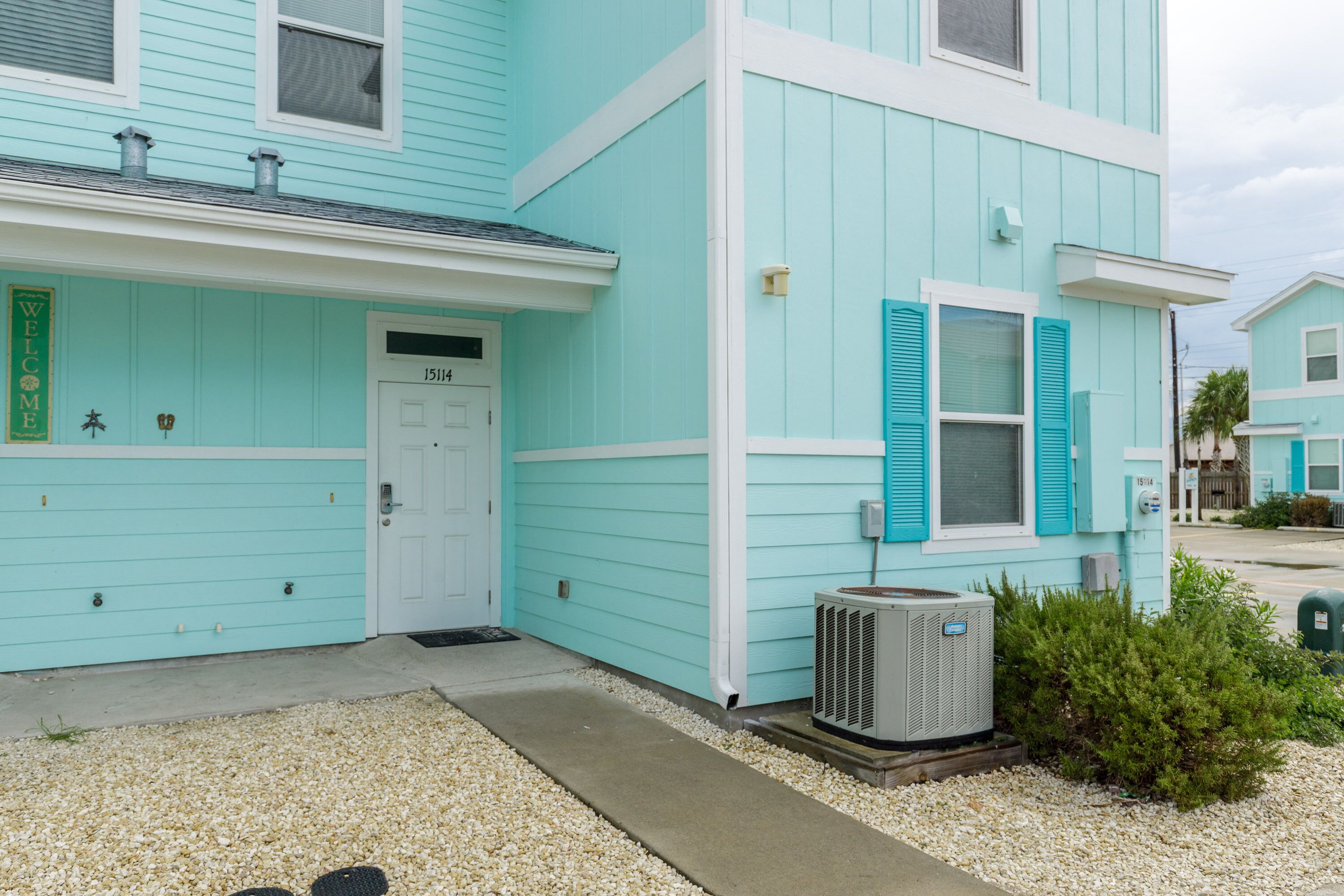 Un bâtiment turquoise à deux étages, avec une porte blanche, un petit climatiseur et une allée gravillonnée.