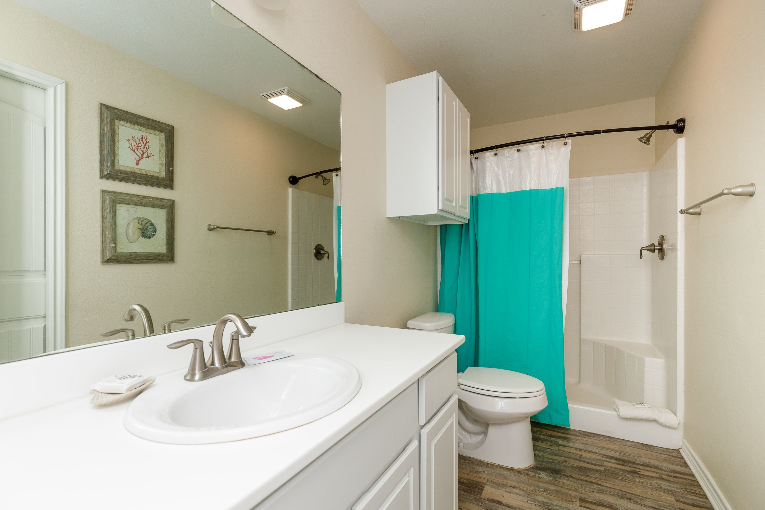 A bathroom with a white sink, toilet, and shower. There are framed pictures on the wall and a white cabinet above the sink.