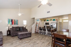 A spacious living room with a gray sofa, ottoman, and a dining area with wooden chairs and a table. A kitchen is visible in the background with a microwave and refrigerator.