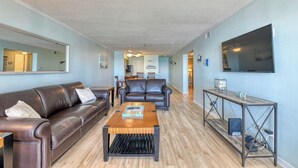 A living room with a brown leather sofa, a wooden coffee table, and a TV mounted on the wall. - Seasider 302 - Two-bedroom, Two & 1/2 Bath Oceanfront Condo (Garden City Beach)