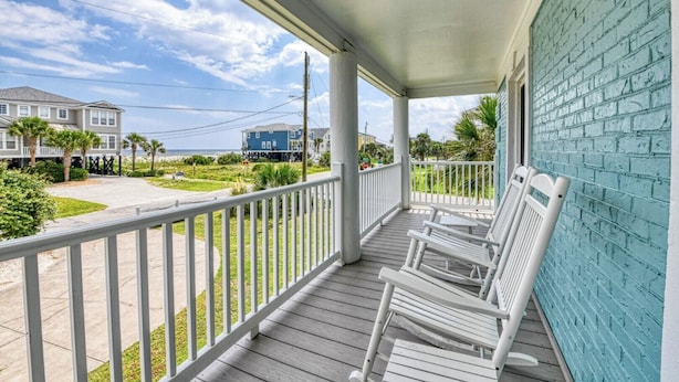 A covered porch with white railings and wooden flooring, overlooking a street with houses and a beach.