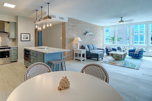 A modern kitchen with a central island, a dining area with a white table, and a living room with a blue sofa and a coffee table.