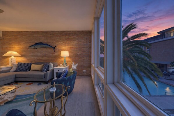 A living room with a brick wall, a sofa, a glass coffee table, and a view of a pool at sunset.