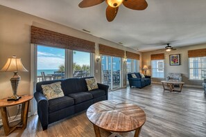A spacious living area with a large window overlooking the ocean, a ceiling fan, and a wooden coffee table. - T's Treasure's - Eight-bedroom, eight-bath oceanfront duplex w/ pool (Surfside Beach)