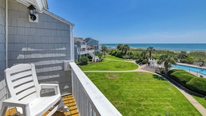 A balcony with a white chair, a pool, and a view of the ocean and beach.