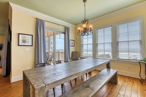 Uma sala de jantar com uma grande mesa de madeira, cadeiras, um candelabro e janelas amplas com persianas.