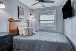 Un dormitorio con cama, mesita de noche, ventilador de techo, ventana con persianas y televisor montado en la pared.