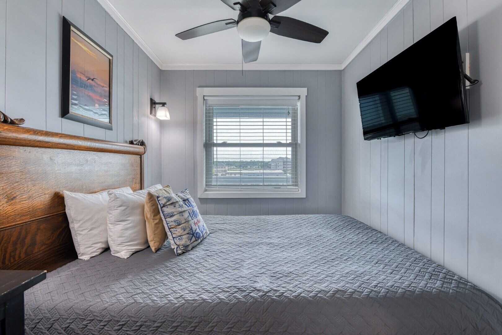 Une chambre avec un lit, un ventilateur de plafond, une fenêtre avec des stores et un téléviseur à écran plat fixé au mur.