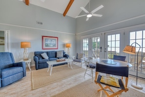 A spacious living room with a blue sofa, a coffee table, and a dining table with chairs. There is a ceiling fan, a framed picture on the wall, and a view through large glass doors.