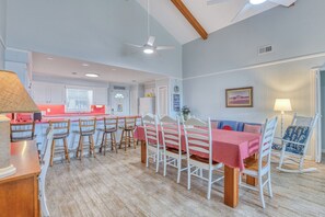 A dining area with a table set for four, surrounded by wooden chairs, a kitchen in the background, and a ceiling fan.